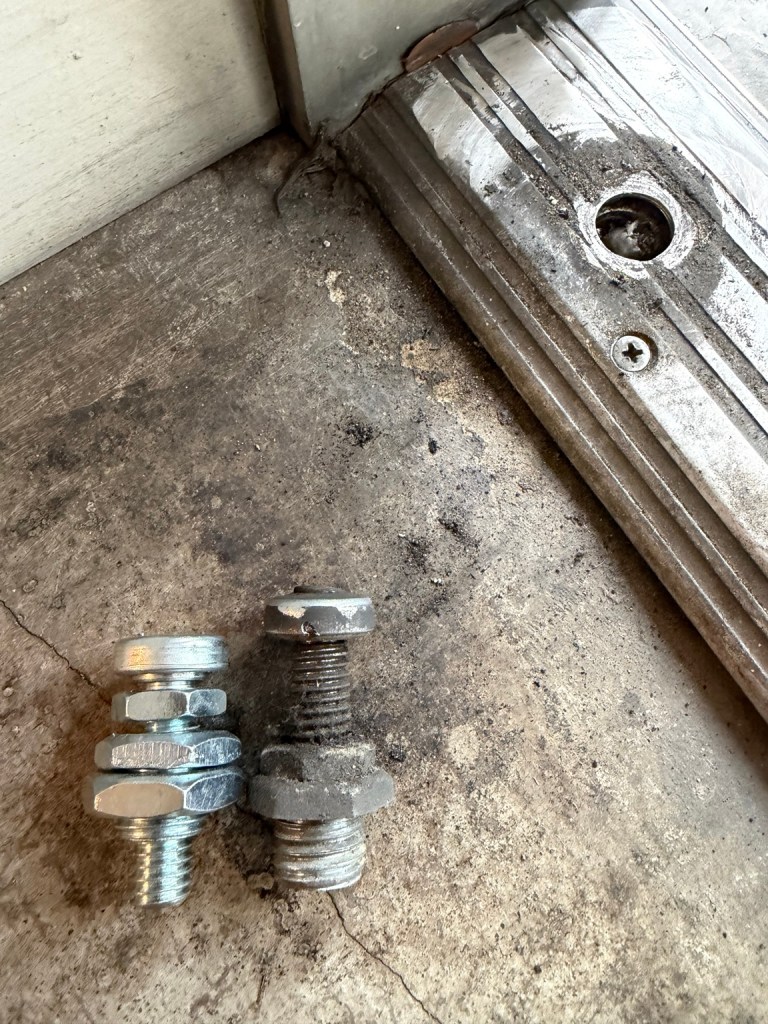 Replacement and existing threaded door pivot pins beside a storefront threshold.
